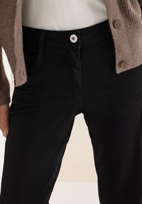 Jeans negros de talle alto con una textura suave, con un botón plateado y trabillas para cinturón, combinados con un cárdigan beige de punto.