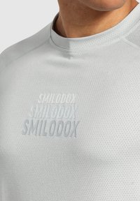 Jasnoszary sportowy t-shirt z oddychającej, teksturowanej tkaniny. Posiada wytłoczone logo "SMILODOX" w różnych rozmiarach czcionek oraz brak widocznych akcesoriów.