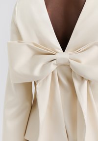 Kremfarget satintopp med dyp V-hals og stor sløyfe bak, med flytende stoff og en jevn tekstur.