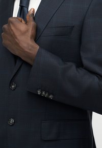 Main d'homme ajustant une veste de costume à carreaux bleu foncé sur une chemise blanche et une cravate sombre, gros plan sur les boutons et la poche.