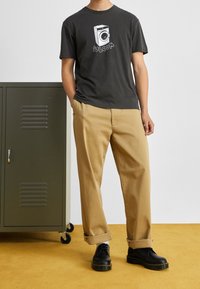 T-shirt grafica grigia con design di una lavatrice, abbinata a chinos beige. Scarpe nere stringate, in piedi accanto a un armadio verde.