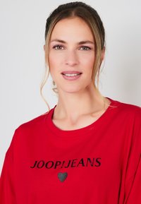 Czerwona koszulka z materiału z czarnym nadrukiem, na którym znajduje się napis "JOOP!JEANS" oraz mały symbol serca pod nim. Miękka tekstura, okrągły dekolt.