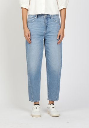 Person wearing light blue high-waist straight-leg jeans with rolled cuffs, white sneakers, and a white shirt, standing against a plain background.