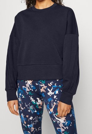 Vrouw die een donkermarineblauwe lange mouwen sweatshirt draagt met geplooide mouwen en kleurrijke abstracte bloemenleggings.
