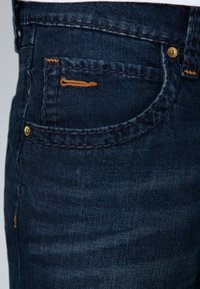 Jeans de mezclilla azul oscuro con costuras en contraste de color naranja, un bolsillo frontal con un diseño distintivo y detalles metálicos en tono dorado.
