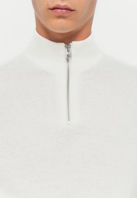 Pull blanc à col montant zippé en tissu tricot doux. Doté d'une fermeture éclair en métal avec un tirette rectangulaire.