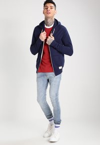 Marineblauwe rits hoodie met witte trekkoorden, gelaagd over een rode shirt, gecombineerd met lichtblauwe jeans en witte sneakers.