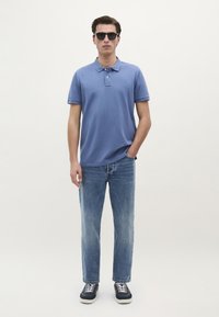 Polo di colore azzurro chiaro con una chiusura a tre bottoni, indossato con jeans dritti blu sbiaditi e sneakers nere con lacci bianchi.