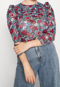Floral blouse with puff sleeves, featuring a blue and red pattern on a sheer material. Paired with high-waisted blue denim jeans.
