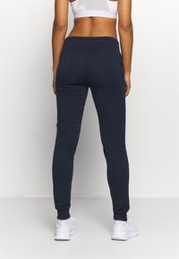 Mujer con joggers azul marino, sujetador deportivo blanco y zapatillas blancas, de pie de espaldas sobre un fondo blanco liso.