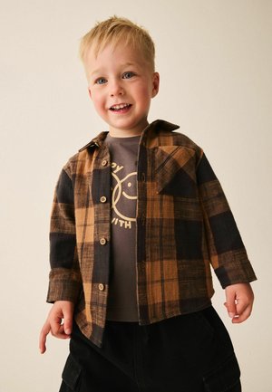 Smilende ung dreng med blondt hår iført en brun og sort ternet skjorte over en grafisk t-shirt og sorte bukser mod en ensfarvet baggrund.