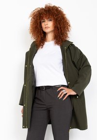 Giacca impermeabile verde oliva con cappuccio, dotata di accessori dorati e tasche laterali, indossata sopra una maglietta bianca e pantaloni grigio scuro.