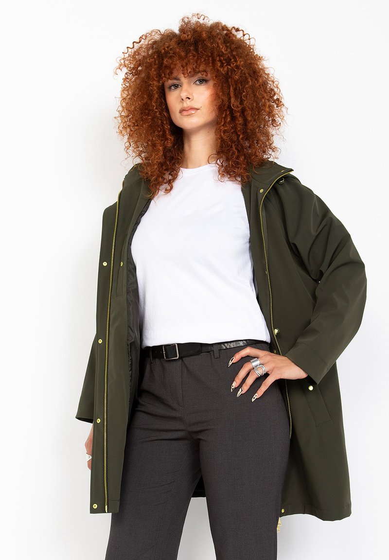 Giacca impermeabile verde oliva con cappuccio, dotata di accessori dorati e tasche laterali, indossata sopra una maglietta bianca e pantaloni grigio scuro.