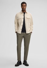 Beige corduroy shirtjack met twee borstzakken, daaronder een zwart t-shirt, olijfgroene op maat gemaakte broek, en zwarte veterschoenen.
