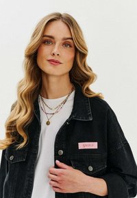 Czarna kurtka dżinsowa z różową łatą "Girly", srebrnymi guzikami i luźnym krojem, połączona z warstwowanymi naszyjnikami na białej koszuli.