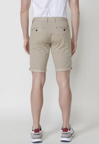 Beige shorts with a rolled hem, two back pockets with button closures, and a flat front design, made from a lightweight fabric.