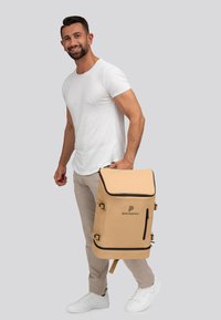 Zaino beige realizzato in materiale resistente, con dettagli neri, logo, cerniera laterale e spallacci regolabili. Progettato per la portabilità.