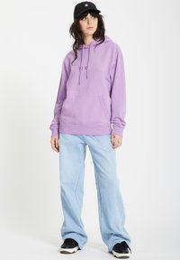 Jasnofioletowa bluza z kapturem z regulowanym sznurkiem, kieszenią kangurką i ściągaczami. Połączona z luźnymi jasnoniebieskimi dżinsami i czarnymi sneakersami.