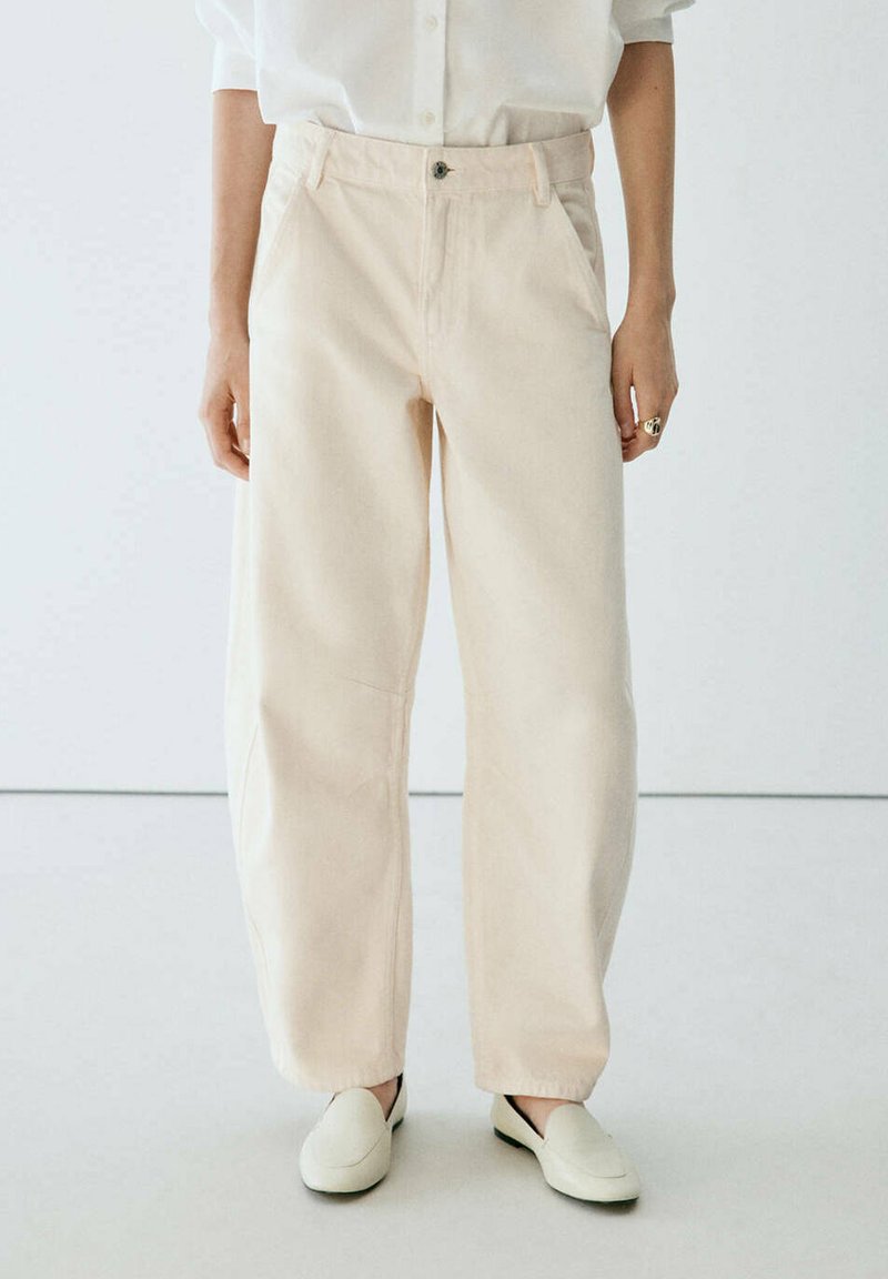 Personne portant un pantalon beige ample, une chemise blanche boutonnée et des chaussures beige assorties sans lacets, debout devant un fond blanc uni.