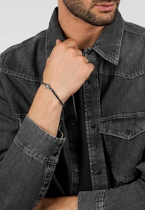 Uomo che indossa una giacca di jeans nera con un braccialetto nero di perline adornato da un piccolo ciondolo d'argento al polso vicino al mento.