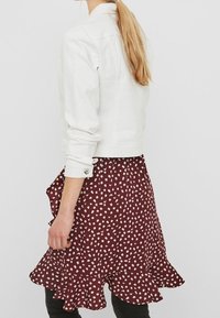 Kvinna med långt blont hår, iklädd en vit jacka och en röd kjol med prickar och volanger, stående mot en enfärgad bakgrund.