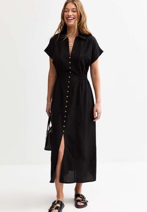 Schwarzes Leinen-Knopfkleid mit kurzen Ärmeln, elastischem Bund, Schlitz vorne und Holzknöpfen. Kombiniert mit schwarzen Riemchensandalen und einer kleinen Tasche.