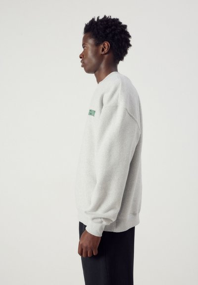 Jeune personne aux cheveux courts et bouclés, portant un sweat-shirt gris clair et un pantalon noir, debout de profil sur un fond blanc uni.