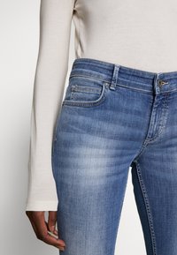 Los jeans de denim azul presentan un clásico diseño de cinco bolsillos, con un acabado desgastado y una cintura de altura media. La tela muestra una textura suave.