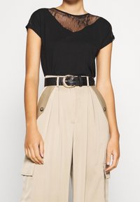 Svart kortärmad topp med en genomskinlig spets framtill; beige högmidjade byxor med veck och sidofickor; svart krokodilpranat bälte med guldspänne.