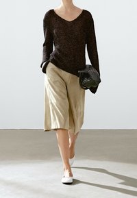 Personne marchant à l'intérieur portant un pull en maille foncée, un short beige jusqu'aux genoux, des chaussures blanches et tenant un sac noir tressé.