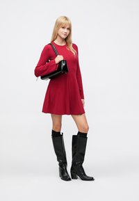 Piros, hosszú ujjú ruha bővülő szegéllyel, térdig érő fekete bőr csizmával és egy kis fekete kézitáskával, egyszínű háttér előtt.