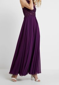 Fioletowa sukienka do ziemi z koronkowym górnym partem ozdobionym koralikami oraz zwiewnym dołem z szyfonu; cechuje się halterowym dekoltem i subtelnym blaskiem.