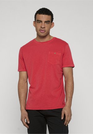 Jeune homme aux cheveux courts et bouclés portant une chemise rouge à manches courtes avec une poche poitrine et un pantalon noir, se tenant devant un fond uni.