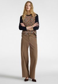 Frau mit langen blonden Haaren, die ein gemustertes Langarmoberteil, weite braune Hose und dunkle spitz zulaufende Absätze trägt, steht mit verschränkten Armen.