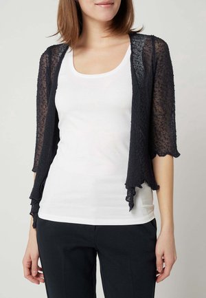 Femme portant un top blanc sans manches sous un cardigan noir transparent à manches courtes et un pantalon noir, debout devant un fond uni.