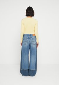 Ανοιχτόκίτρινη μπλούζα με μακριά μανίκια, συνδυασμένη με μπλε τζιν παντελόνι με φαρδιά πόδια, διαθέτοντας αντίθετο κάτω πάνελ, πίσω τσέπες και ετικέτα μάρκας στη μέση.