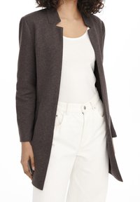 Blazer largo marrón oscuro con cuello de muesca, textura suave y mangas ajustadas, que se lleva sobre una camisola blanca y pantalones blancos.