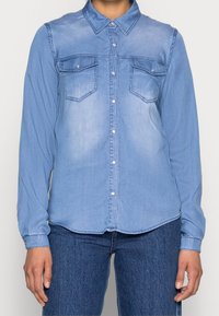 Light blue denim shirt with long sleeves, standard collar, two front pockets, and button closure. Soft texture with a slightly faded finish.