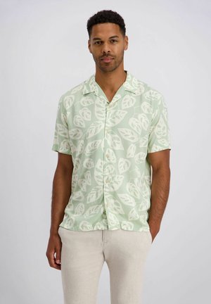 Man draagt een lichtgroen overhemd met korte mouwen en een wit bladpatroon, gecombineerd met beige broek, staand tegen een effen achtergrond.