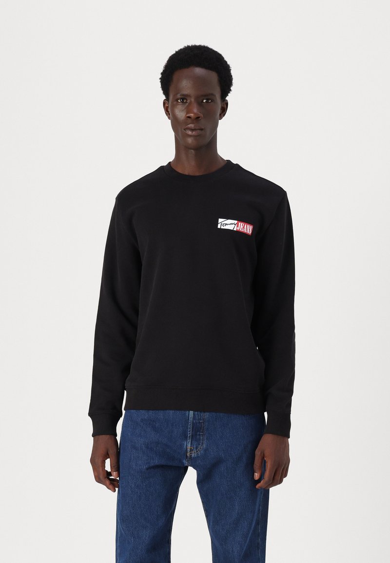 Svart hettegenser med et lite rødt og hvitt logo på brystet. Stoffet ser mykt ut, med rund hals og ribbestrikkede ermer.