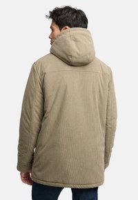 Olivově zelená manšestrová bunda s velkou kapucí, žebrovanou texturou a detaily na předním švu, doplněná tmavými džínovými kalhotami.