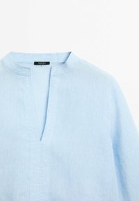 Vaaleansininen pellavapaita, jossa on bändikaulus ja V-muotoinen aukileikkaus, jossa on hienovaraisia ommeltuja yksityiskohtia ja pieni musta etiketti kauluksessa.