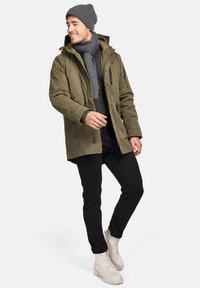 Oliven grøn parka med lynlåslukning og knaplukning, grå strikket tørklæde, matchende beanie, sorte bukser og lyse støvler med snøringer.