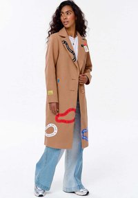 Žena s dlouhými kudrnatými vlasy má na sobě béžový kabát s barevnými nášivkami s nápisem "NEZASTAVUJ", světle modré džíny, bílé tenisky a vrstvené náhrdelníky.