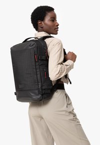 Svart ryggsäck med en texturerad design och röda accenter, flera blixtlåsfickor och justerbara remmar, bärs på ena axeln.