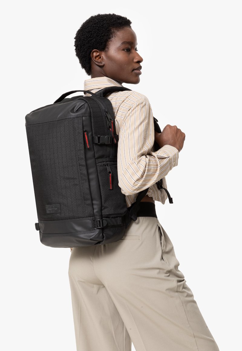 Svart ryggsäck med en texturerad design och röda accenter, flera blixtlåsfickor och justerbara remmar, bärs på ena axeln.