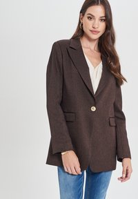 Blazer marrone con texture e vestibilità su misura, un bottone dorato, revers a battente e due tasche frontali, abbinato a una maglietta chiara e jeans.