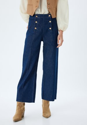 Pantaloni in denim a gamba larga di un blu scuro, con vita alta e quattro grandi bottoni circolari sulla parte anteriore. Il tessuto sembra robusto con una superficie liscia.