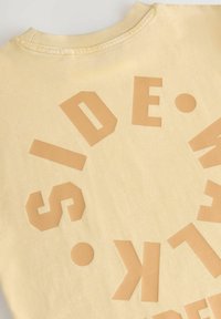 Sweatshirt en coton jaune clair avec un motif circulaire comportant des lettres et des formes en cuir tan et en gras. Col côtelé et texture douce.