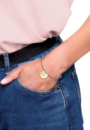 Bracciale color oro con pendente circolare inciso "migliore mamma", attaccato a un sottile cinturino grigio. Indossato al polso, abbinato a jeans e una camicia leggera.
